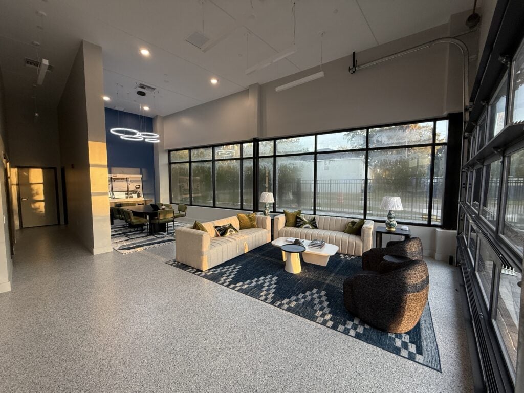 Modern office lounge with large windows, beige sofas, a dark armchair, and a blue patterned rug, featuring contemporary decor and natural lighting.