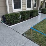 Newly coated concrete driveway and walkway with a landscaped garden bed in front of a house.