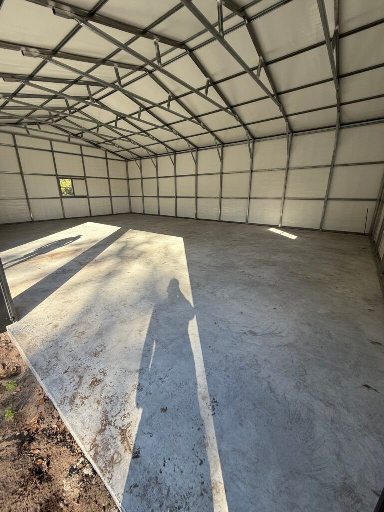 Empty garage with concrete floor under a metal roof, illuminated by sunlight.