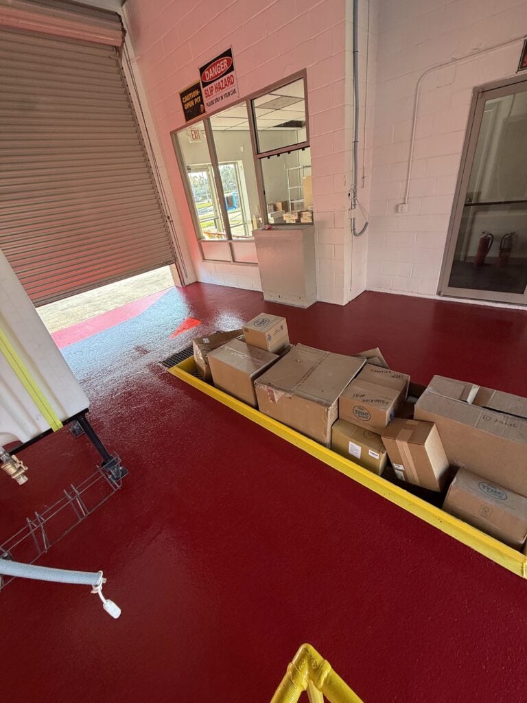 Boxes of supplies inside Checkmate Coatings facility with red flooring and large garage door.