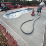 Pool deck construction with concrete surface and a partially installed pool, workers applying coating, and construction equipment present.