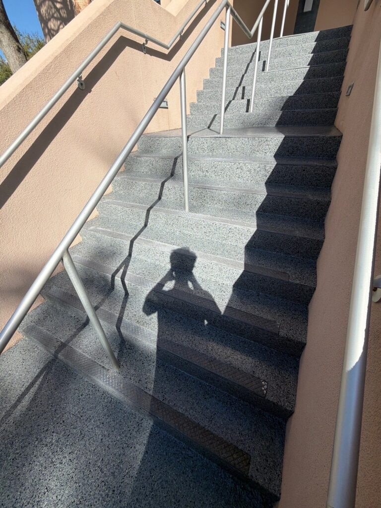 Coated Concrete staircase with metal handrails outside a building, captured during daytime with shadows and sunlight.